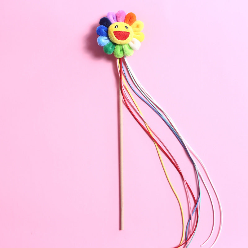 Bâton Teaser en tissu avec grelots et pompons multicolores pour chat, forme de fleur souriante sur fond rose.