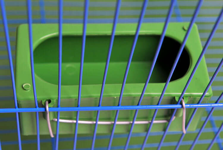 Conteneur de nourriture pour oiseaux étanche fixé à une cage avec barreaux bleus.