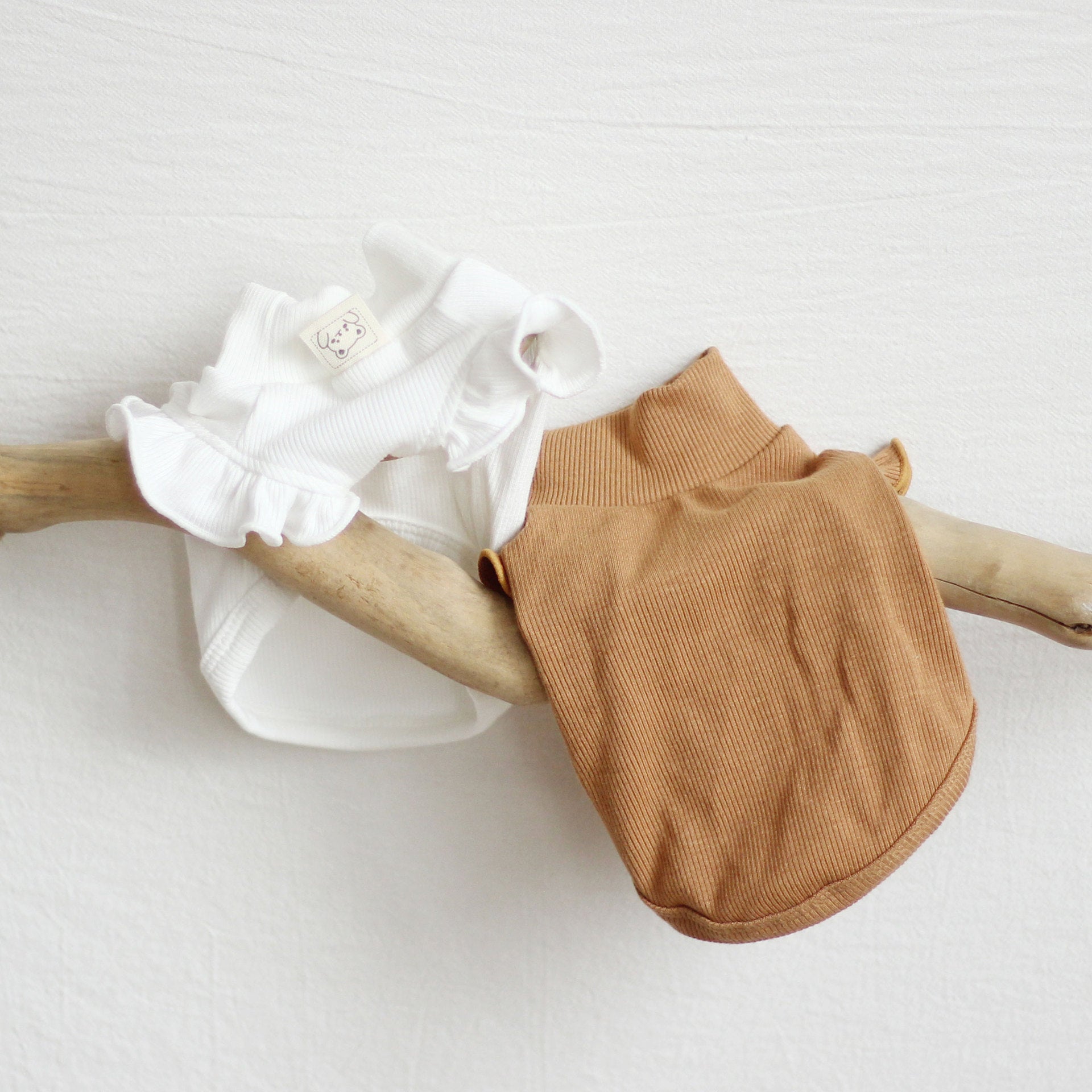 Deux vêtements pour animaux, un haut blanc et un pull brun, accrochés sur une branche en bois.