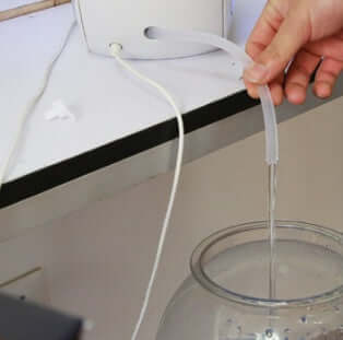 Person filling an AquaDesign aquarium with water using a clear hose.
