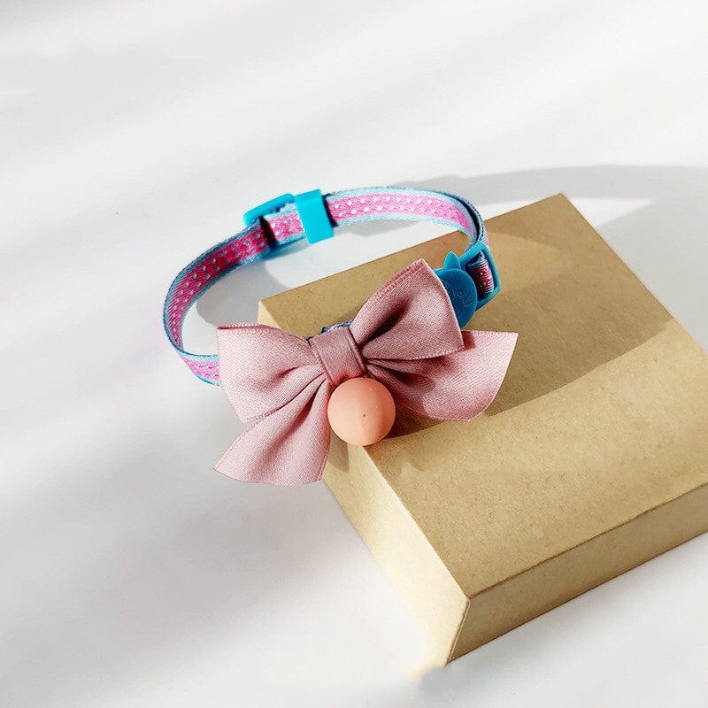 Nœud papillon rose pour chat avec cloche sur collier bleu, présentée sur une boîte marron.