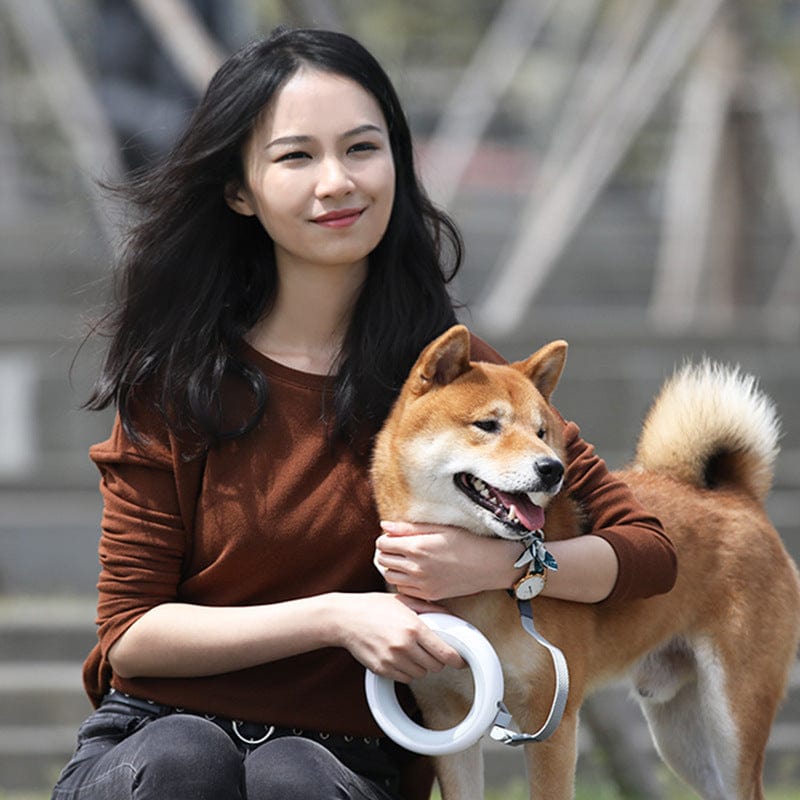 Femme souriante tenant une laisse rétractable avec un chien Shiba Inu lors d'une promenade.