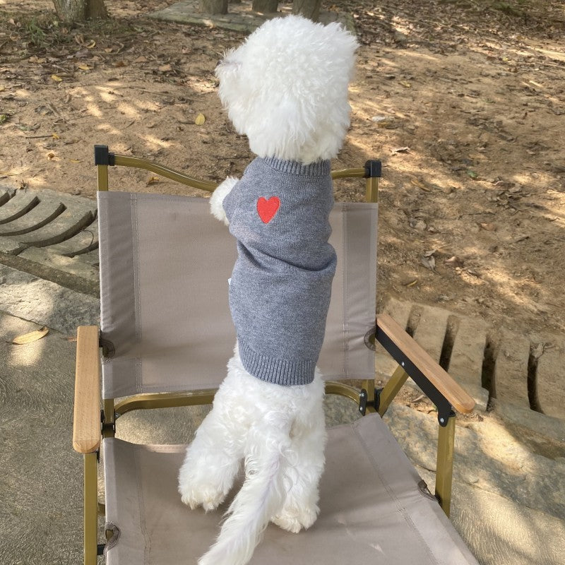Chien en pull en laine gris avec cœur rouge, assis sur une chaise en plein air.